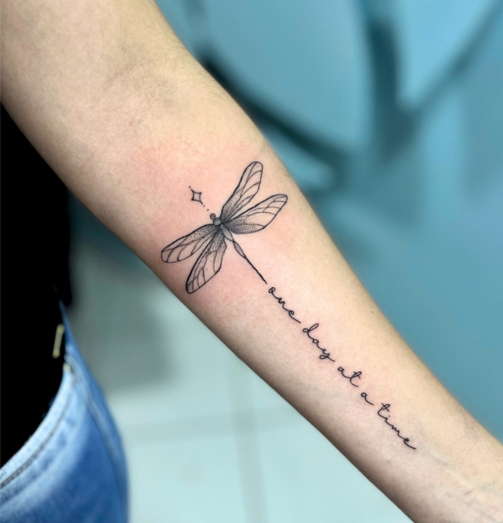 Tatuagem fineline de libélula com lettering no antebraço